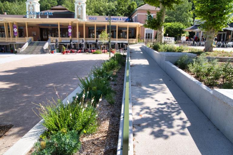 Ax-les-Thermes : Aménagement paysager et urbain devant un casino: cheminement piéton en béton désactivé bordé de massifs végétalisés, terrasses animées en arrière-plan sous une architecture patrimoniale.