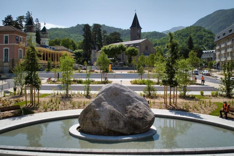 Ax-les-Thermes : Place urbaine paysagère de montagne : fontaine circulaire avec rocher central, parterres plantés et jeunes arbres alignés, pavages clairs, église en arrière‑plan et bâtiments historiques autour.