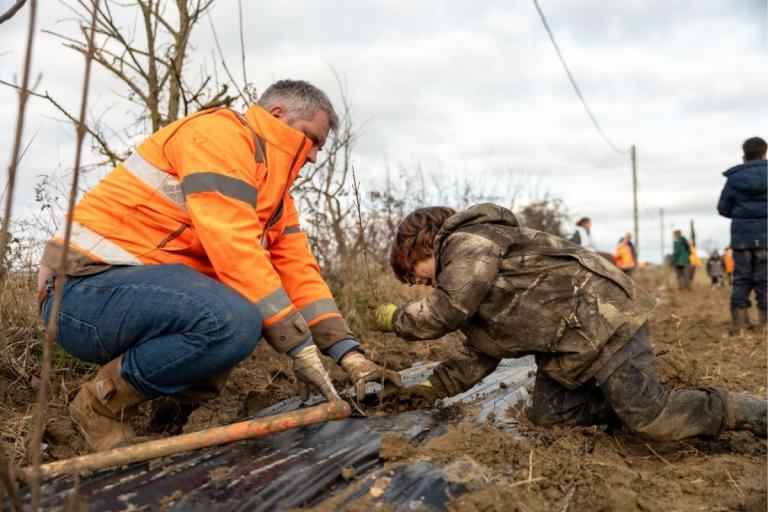 Chantier de plantation participative : encadrant en gilet orange accompagne un enfant qui met en terre un jeune plant à travers un film de paillage, sol très boueux, rang planté le long d’une haie.