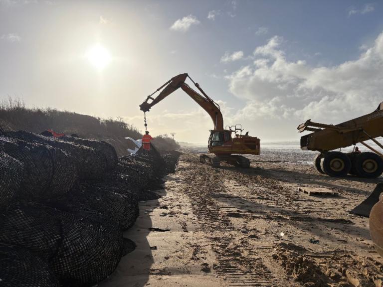 Oléron face à l'érosion : Travaux de protection côtière : pelle hydraulique manutentionnant de gros rouleaux/structures en géonets sur une plage, engins de chantier sur le sable et mer en arrière‑plan sous un soleil bas.