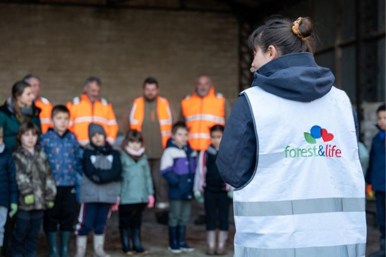 Sensibilisation forestière avec scolaires : animatrice en gilet « forest&life » de dos face à un groupe d’enfants et d’adultes en gilets haute visibilité, scène en intérieur/abri avant sortie terrain.