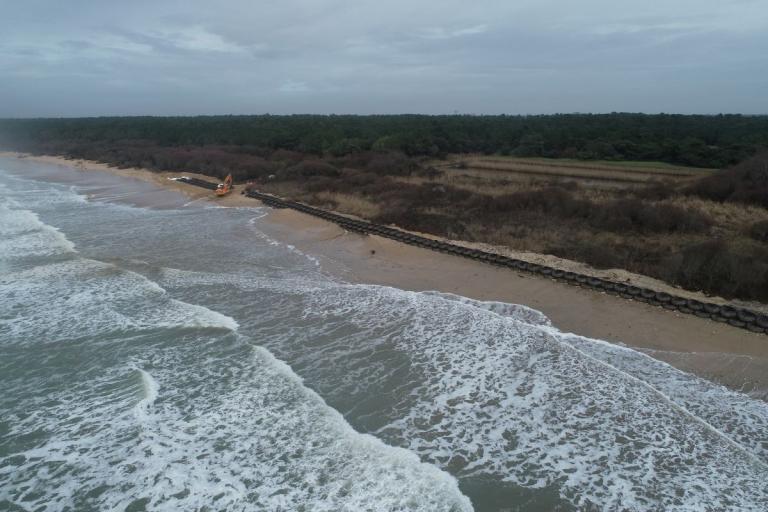Oléron face à l'érosion : Pose de digue souple sur plage atlantique : pelleteuse et équipe installent une longue ligne de boudins/géotubes en géonets le long du haut de plage, houle et forêt littorale en arrière‑plan.