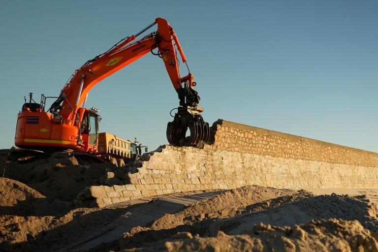 Pelle hydraulique Komatsu équipée d’un grappin remet en état un mur de quai au lever du soleil, avec tombereau en appui et dunes de sable en premier plan.