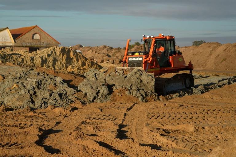 Bulldozer Komatsu orange repousse une levée de déblais au petit matin, sculptant la plateforme d’un futur chantier entre merlons de sable et bâtisse en lisière.