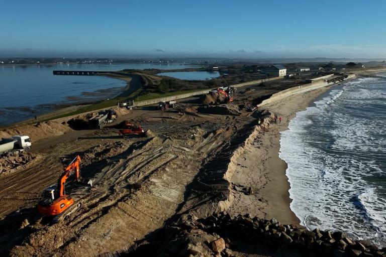 Chantier de lutte contre l’érosion littorale à Oléron : pelle hydraulique posant des boudins/géotextiles en file le long d’une dune protégée par un géotextile blanc, équipe en gilets haute visibilité, plage atlantique et mer à droite.