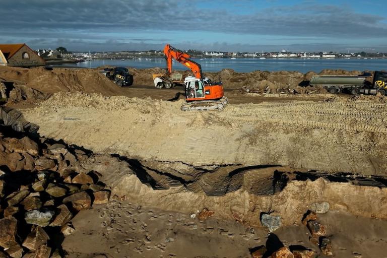 Reprofilage et rechargement de plage/cordon dunaire en zone portuaire : pelle hydraulique orange façonne les volumes de sable, camions bennes en évacuation/apport, ganivelles et enrochements au premier plan, plan d’eau et bourg en arrière‑plan.