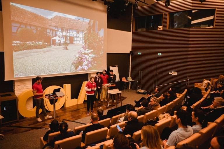 Présentation Challenge Tic t'ACT : Présentation TIC & ACT dans un amphithéâtre Colas : équipe en t‑shirts rouges exposant un projet devant un public, grande projection d’une image de maison à colombages en arrière‑plan.