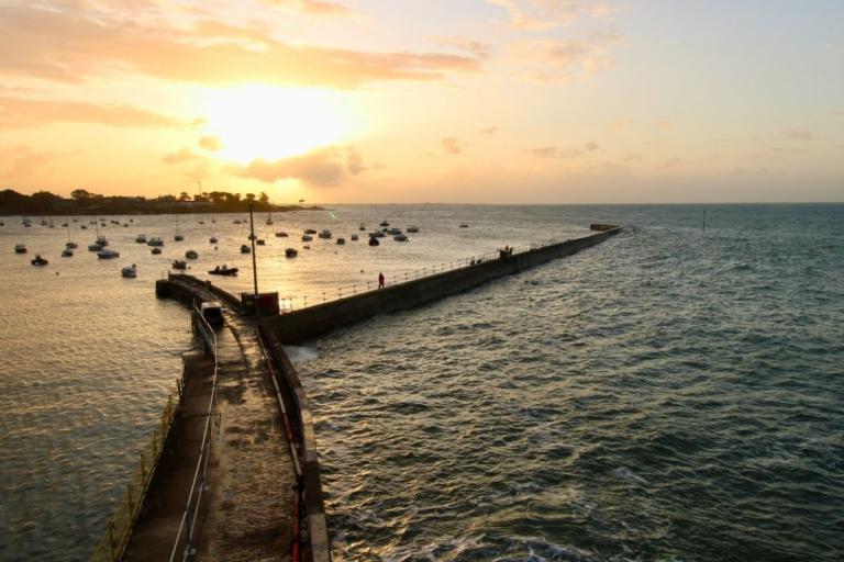 Jetée au coucher du soleil avec mer agitée et nombreux mouillages, accès protégé par barrières le long de l’ouvrage.