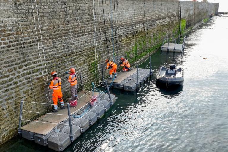 Ouvriers en EPI travaillant le long d’un môle : plateformes flottantes, plongeur en intervention et matériel de chantier alignés le long de l’ouvrage.