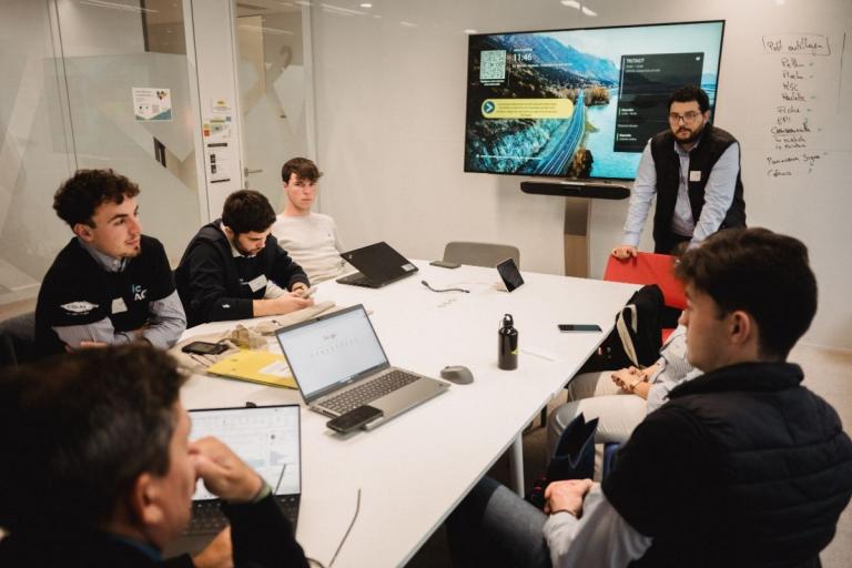 Challenge Tic t'ACT Groupe de travail : Réunion projet en salle de créativité : équipe autour d’une table avec laptops, écran affichant un dashboard TIC&ACT, tableau blanc listant « Pitch », « REX », « Risques », « Planning », « Budget », « EPI », « Communication ».