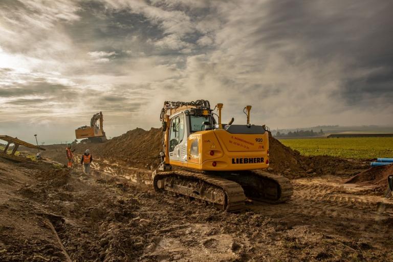 Pelleteuse Liebherr et équipe en EPI sur chantier de tranchée en milieu rural, terrassement et pose de canalisation bleue.
