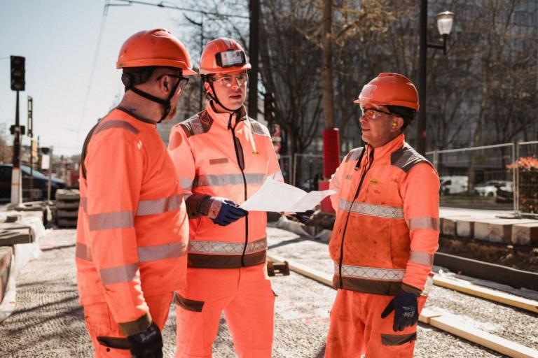Trois collaborateurs de chantier en tenue haute visibilité consultent un plan de travaux au milieu d'un chantier urbain.