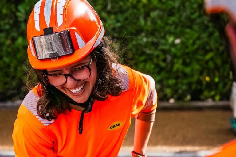 Jeune femme portant un casque et des lunettes sourit pendant une activité sur chantier, dans un environnement extérieur verdoyant.