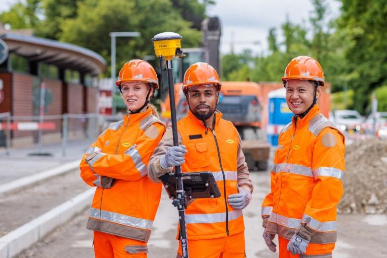 Trois collaborateurs Colas en tenue de chantier orange, dont l’un utilise un équipement de mesure topographique.