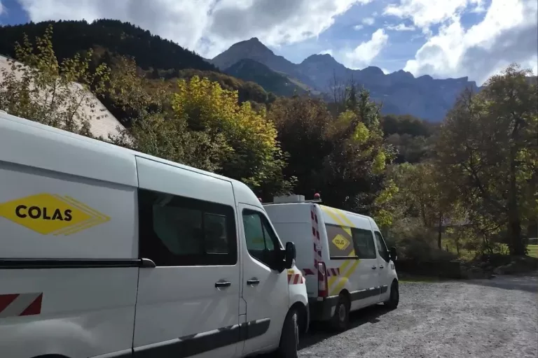 Deux véhicules de chantier Colas stationnés au pied d’un massif montagneux boisé.