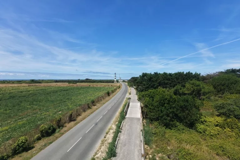 Vue aérienne d’une route et d’un cheminement aménagé entre des espaces agricoles et boisés, en direction du littoral.