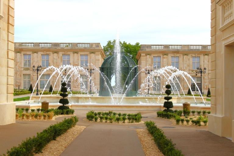 Grande fontaine avec jets d’eau au cœur des jardins du Puy du Fou, entourée de topiaires et de bâtiments d’architecture classique.