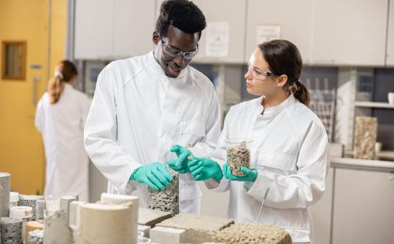 Scientifiques au Core Center : Deux techniciens en blouse blanche dans un laboratoire de matériaux. Ils manipulent des éprouvettes et des granulats de béton sur une table remplie d'échantillons et d'outils de laboratoire ; on voit des contenants, des morceaux de béton et des appareils d'essai en arrière-plan.