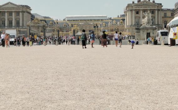 La cour d’entrée du château de Versailles avec ses grilles dorées, des groupes de visiteurs et des cars stationnés sur la droite sous un ciel clair.