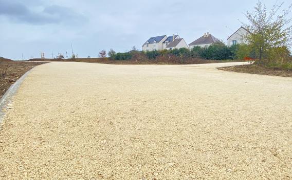 Chemin doux en sable clair serpentant dans un parc résidentiel, reliant les maisons au paysage sous un ciel voilé.