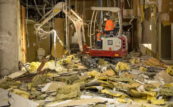 Mini-pelle en action au cœur d’une zone de curage, évacuant gravats, isolants et faux plafonds dans un vaste espace intérieur en démolition.