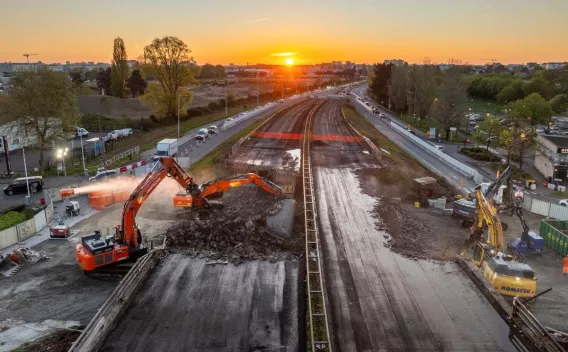 Démolition d’un tablier ferroviaire au crépuscule : deux excavatrices orange attaquent la dalle à gauche, une pelle jaune travaille à droite, poussière en suspension et rails au centre qui s’étirent vers le coucher de soleil.