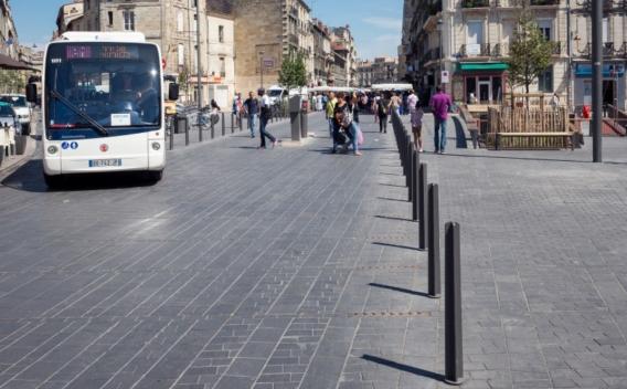 Place piétonne urbaine bordée de commerces avec un bus et des passants circulant entre des bornes alignées.