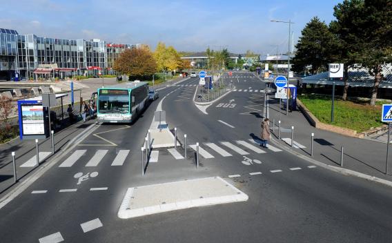 Pôle d’échanges urbain avec voies de bus dédiées, passages piétons, piste cyclable et voyageurs montant à l’arrêt devant des bâtiments modernes.