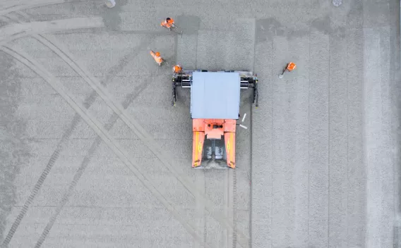Vue aérienne d’une finisseuse orange Colas nivelant un large tapis de granulats, encadrée par quatre opérateurs en gilets haute visibilité.