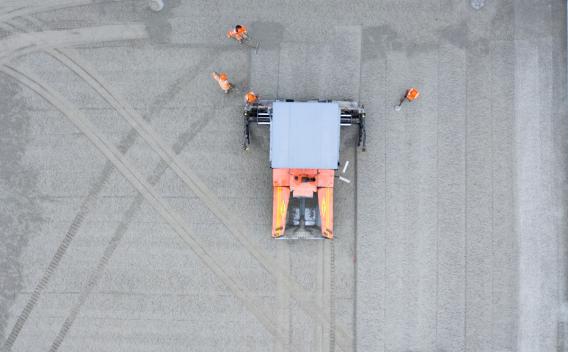 Vue aérienne d’une finisseuse orange Colas nivelant un large tapis de granulats, encadrée par quatre opérateurs en gilets haute visibilité.