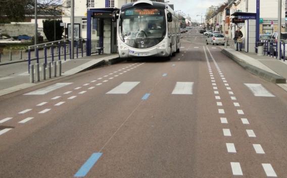 Revêtement de voirie urbain coloré (enrobé massif teinté), marquages longitudinaux et passages piétons appliqués, stationnement et arrêt de bus en site propre. Circulation locale et mobilier urbain visibles.