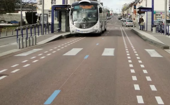 Revêtement de voirie urbain coloré (enrobé massif teinté), marquages longitudinaux et passages piétons appliqués, stationnement et arrêt de bus en site propre. Circulation locale et mobilier urbain visibles.