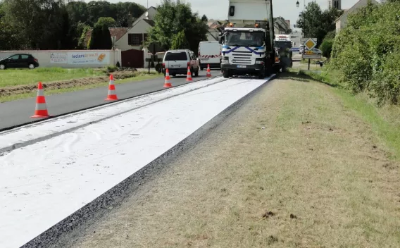 Application d’un géotextile suivi de la pose d’enrobé sur une route en milieu semi-urbain avec balisage de chantier et véhicule d’approvisionnement.