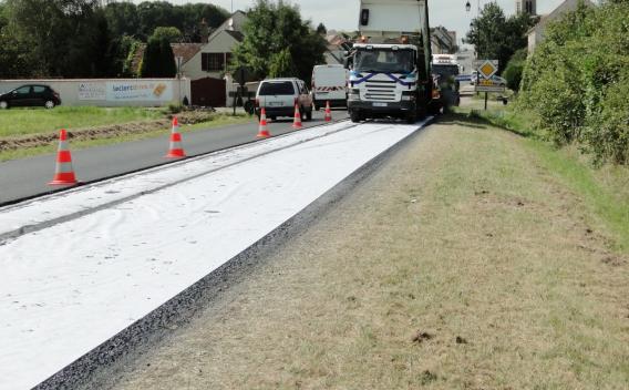 Application d’un géotextile suivi de la pose d’enrobé sur une route en milieu semi-urbain avec balisage de chantier et véhicule d’approvisionnement.