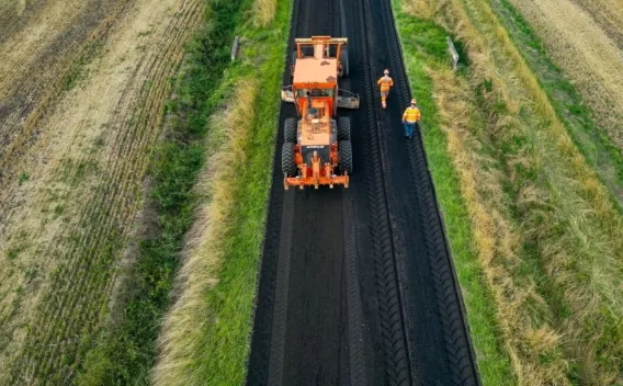 Un finisseur applique une couche d'enrobé sur une route rurale, accompagné d'ouvriers en haute visibilité et de véhicules d'approvisionnement.