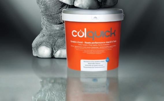 Seau orange de produit "Colquick" (enrobé à froid, haute performance réactif à l’eau) posé sur sol réfléchissant, avec pied d’éléphant en arrière-plan.