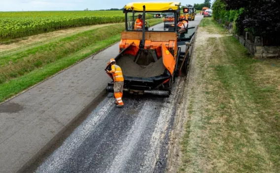 Vue aérienne d’un engin finisseur posant une chaussée au milieu d’un paysage rural, entouré d’ouvriers en tenue de chantier.