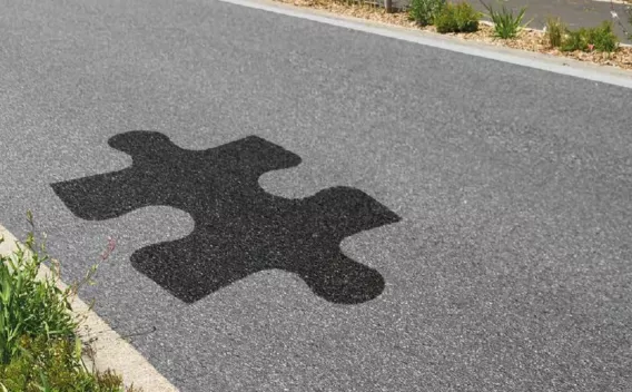 Motif ludique en forme de pièce de puzzle incrusté en enrobé noir sur une chaussée grise bordée de végétation.