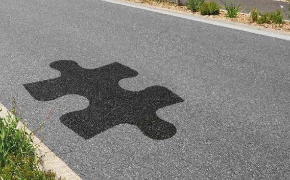 Motif ludique en forme de pièce de puzzle incrusté en enrobé noir sur une chaussée grise bordée de végétation.