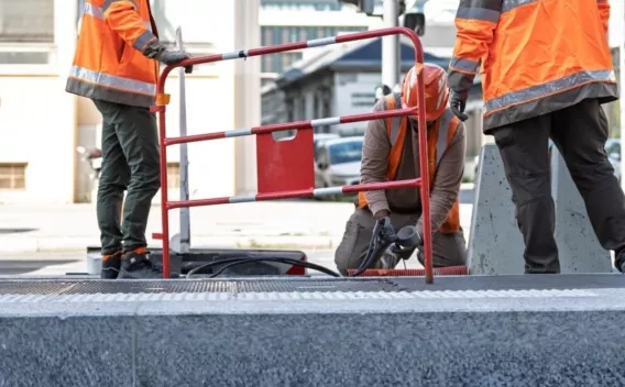 Trois ouvriers en tenue de sécurité travaillent autour d’une ouverture dans le sol, protégée par une barrière de chantier.