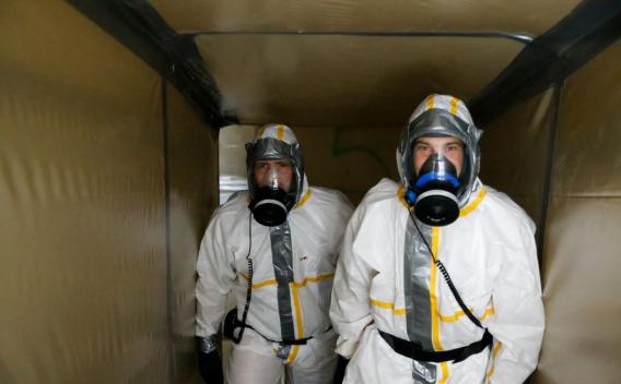 Deux opérateurs en tenue de protection complète (combinaisons blanches avec bandes jaunes, cagoules et appareils respiratoires complets) se tiennent côte à côte dans un sas/confiner de chantier ; ambiance de décontamination ou d’intervention sur matériaux dangereux.