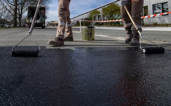  Gros plan sur l’application d’un revêtement noir sur la chaussée au rouleau, avec seau de produit à proximité.
