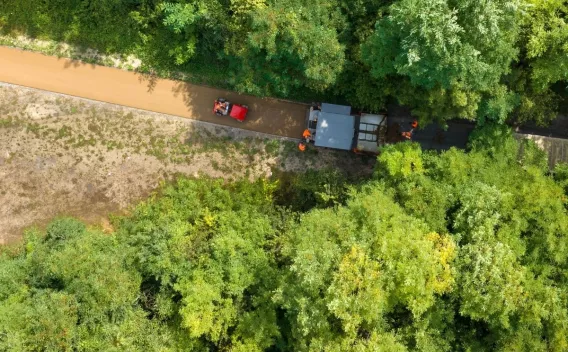 Vue aérienne d’un engin finisseur posant une piste piéton/cyclable claire entourée d’un boisement dense avec plusieurs ouvriers au bord.