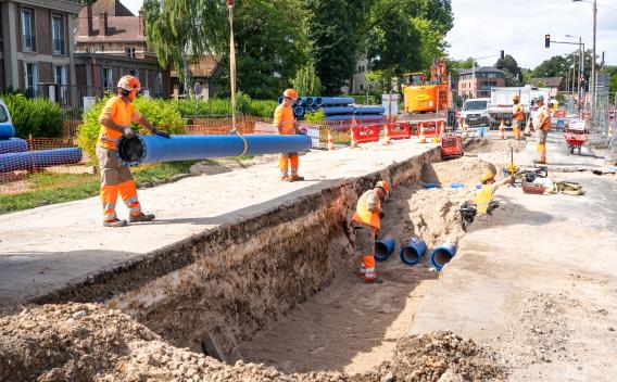 Pose de conduites en PVC bleu dans une tranchée routière : plusieurs opérateurs en tenue haute visibilité coordonnent le positionnement et l'assemblage des sections de tuyau avec engins et barrières de chantier en arrière-plan.