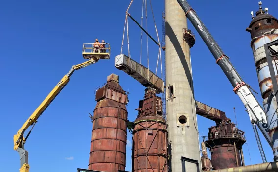 Site industriel ancien en cours d'intervention : grues et nacelles en cours de manutention et de levage autour de grandes colonnes et cuves métalliques rouillées, ciel bleu.
