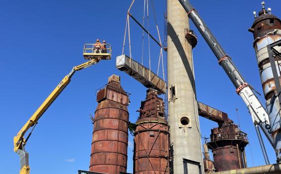 Site industriel ancien en cours d'intervention : grues et nacelles en cours de manutention et de levage autour de grandes colonnes et cuves métalliques rouillées, ciel bleu.