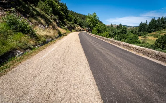 Route de montagne partiellement remise à neuf : une moitié d’enrobé neuf bordée de végétation et d’un talus rocheux, engin de chantier au loin sous un ciel bleu.