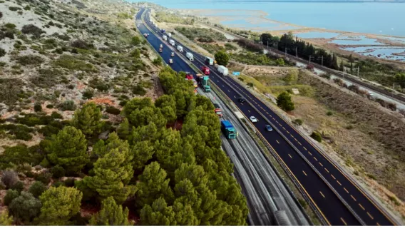 Chantier nocturne sur autoroute en milieu naturel littoral : convoi d’engins et camions sur une chaussée fraîchement refaite, circulation maintenue sur voies adjacentes, voie ferrée et étangs en parallèle.
