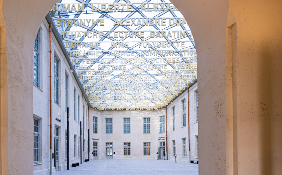 Cour intérieure restaurée d’un bâtiment patrimonial, couverte d’une verrière artistique composée de mots découpés, créant un puits de lumière au-dessus des façades en pierre claire.