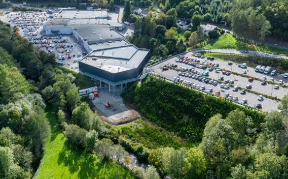 Vue aérienne d’un centre commercial moderne bordé de forêts, avec un vaste parking en pente et une zone en construction.